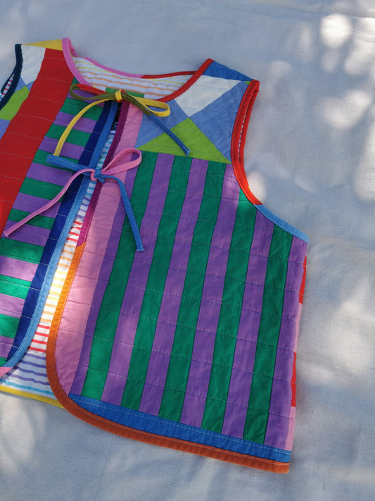 A colourful handmade quilted vest with vertical stripes in green, purple, blue, red, and orange, featuring a tie-neck closure, laid flat on a light fabric background with dappled sunlight by KABIN NZ.