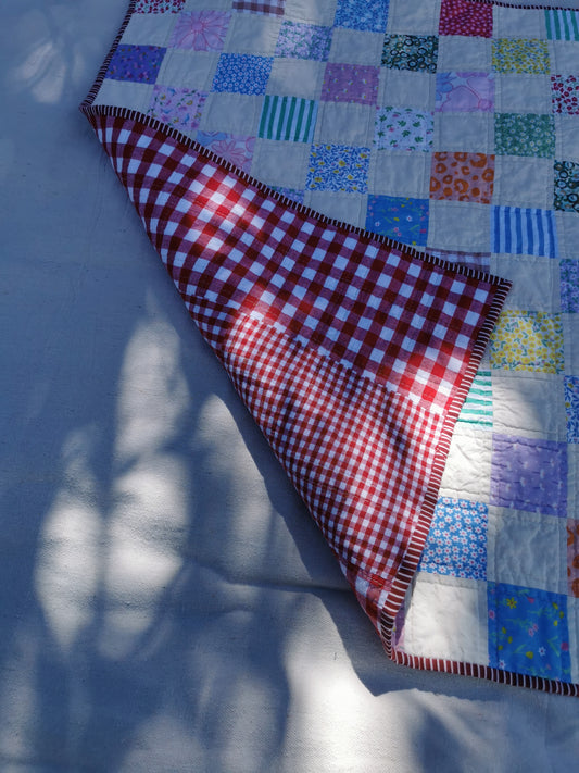 A colourful handmade patchwork quilt with a red and white gingham reverse side, laid out on a white fabric surface with dappled sunlight and shadows by KABIN NZ.