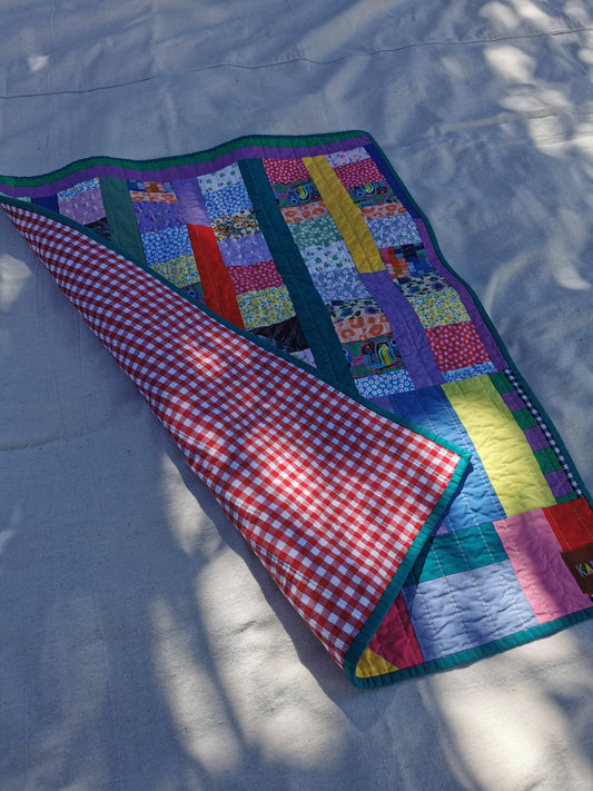 A colorful handmade patchwork quilted blanket with a red and white gingham reverse side, laid out on a light fabric surface with dappled sunlight by KABIN NZ.