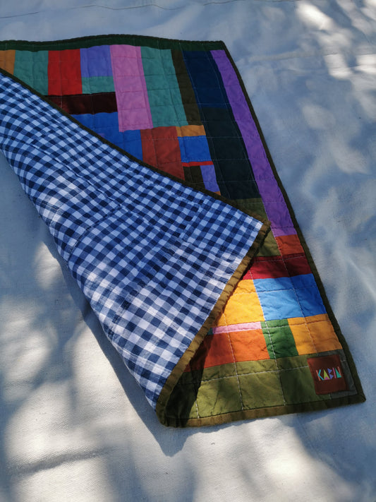 A folded quilted blanket with a multicolor patchwork design and a blue-and-white gingham reverse side, laid on a white surface with dappled sunlight and shadows by KABIN NZ.