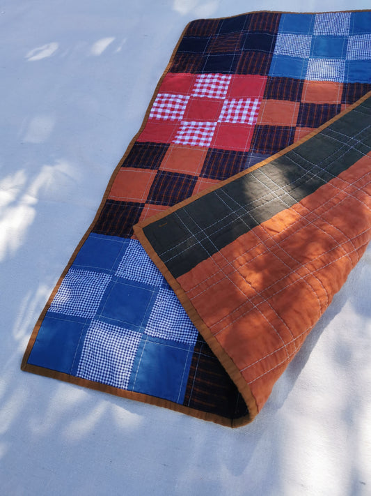 A handmade patchwork quilt laid out on a light surface with dappled sunlight. The quilt features a mix of blue, red, brown, and black checkered and gingham patterns, with a solid brown border by KABIN NZ.