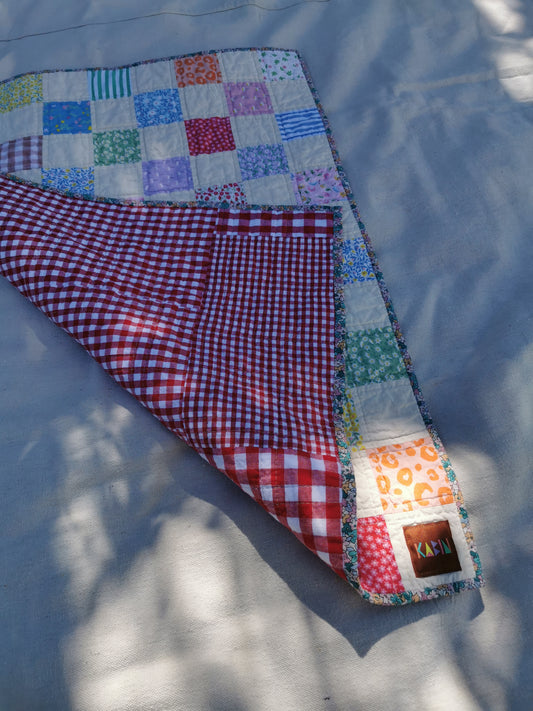 A handmade patchwork quilted blanket with a red and white gingham reverse side and a multicolor patchwork front, laid out on a light gray fabric background with dappled sunlight by KABIN NZ.