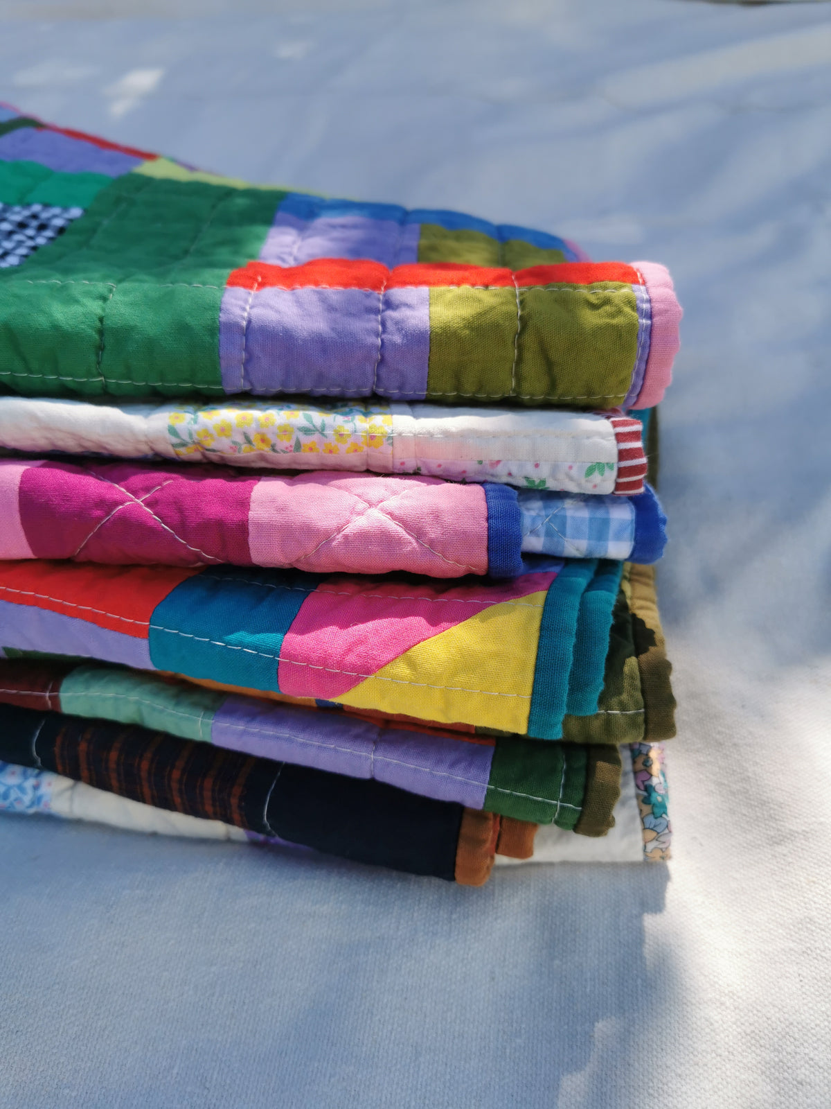 A stack of colourful handmade patchwork quilts resting on a light fabric surface under natural sunlight by KABIN NZ.