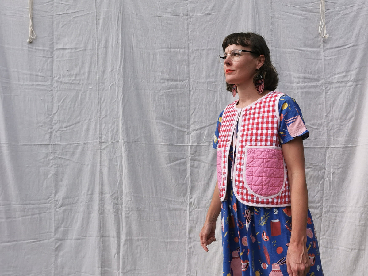 Woman wearing a red and white checkered vest with pink patch pockets over a blue dress with a pattern, standing against a plain background.