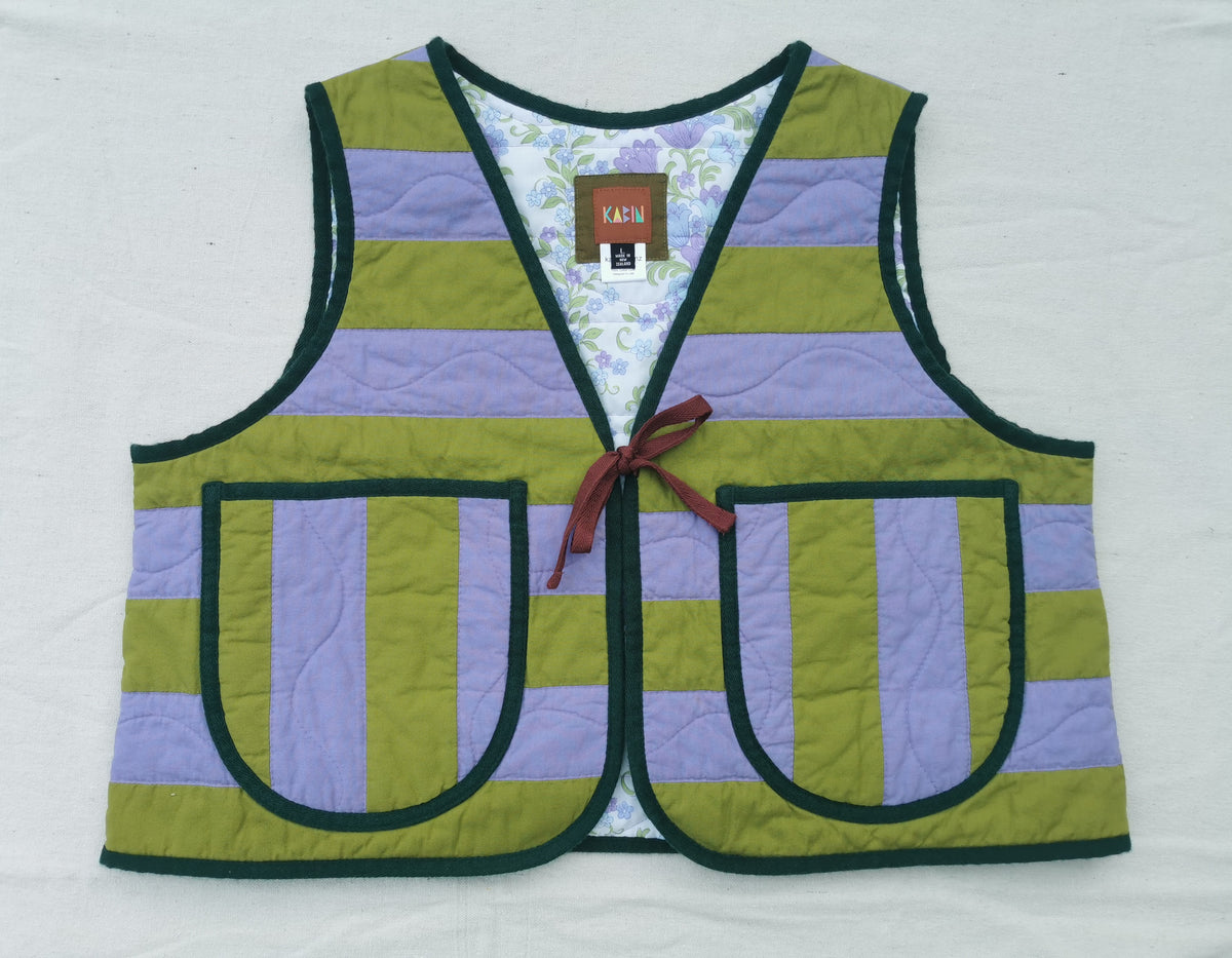 A handmade quilted vest by KABIN NZ with horizontal olive green and lavender purple stripes, dark green trim, two front patch pockets, and a burgundy tie closure, laid flat on a white background.