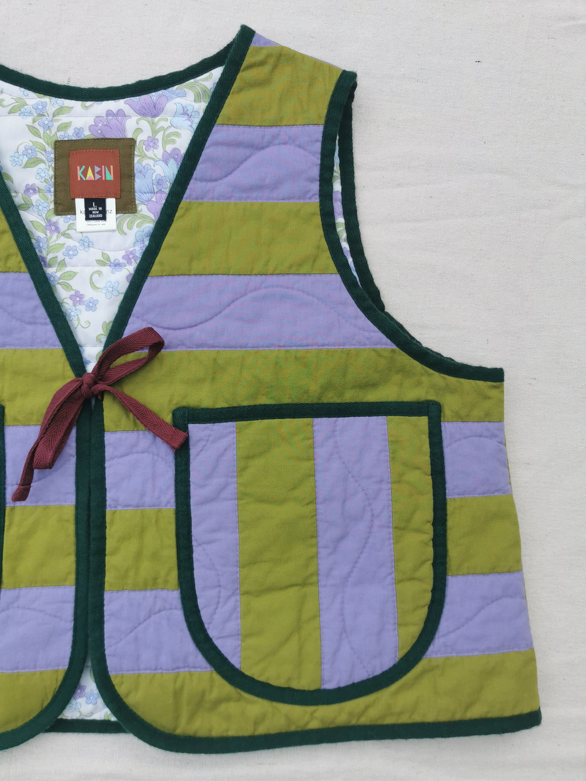 A handmade quilted vest by KABIN with horizontal stripes of olive green and lavender purple, dark green trim, a burgundy tie at the front, and a vintage floral-patterned lining. 