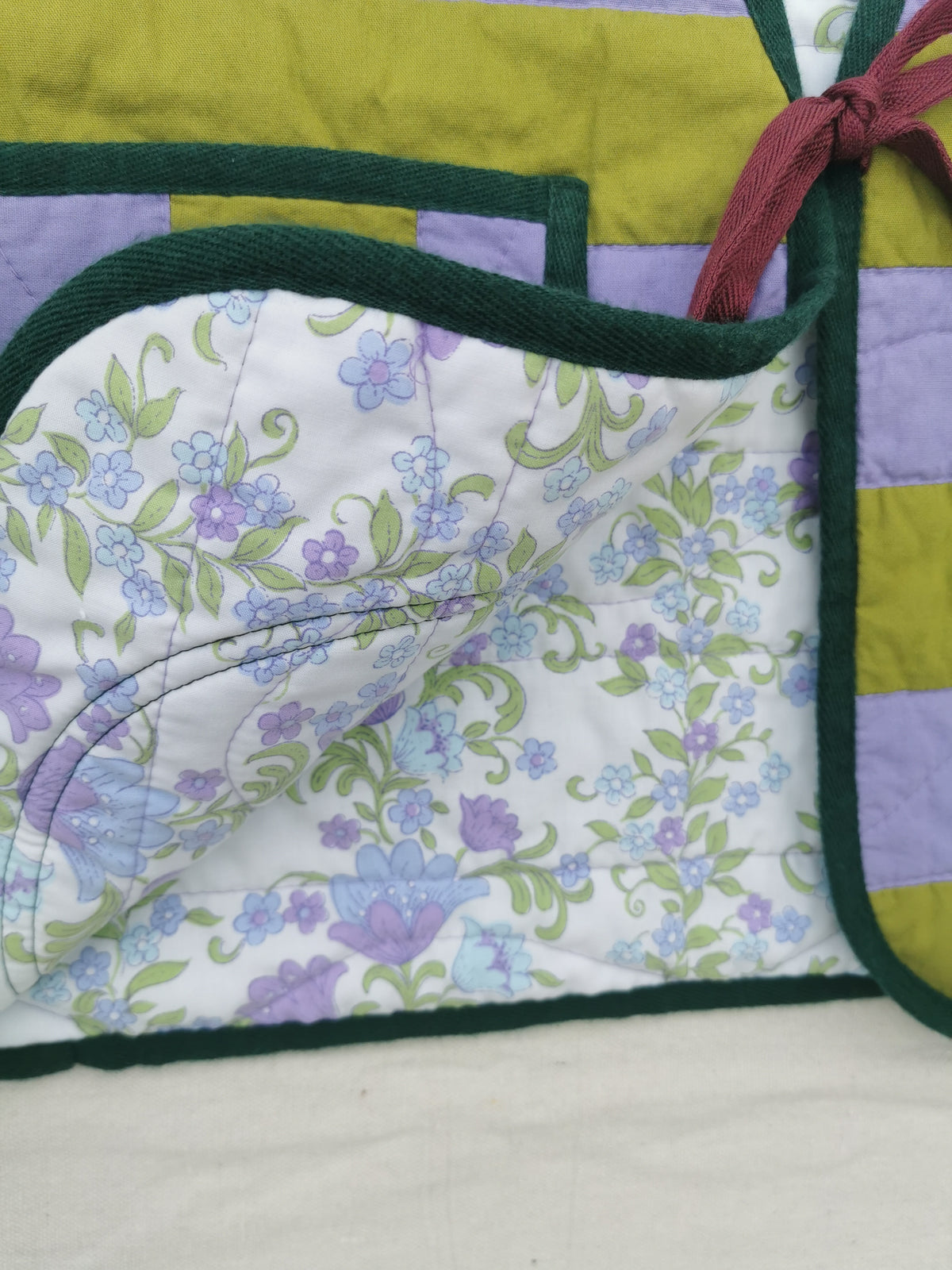 A close-up of a handmade quilted vest by KABIN, showing a floral patterned lining with purple and blue flowers on a white background, dark green borders, and lime green and lavender striped sections.