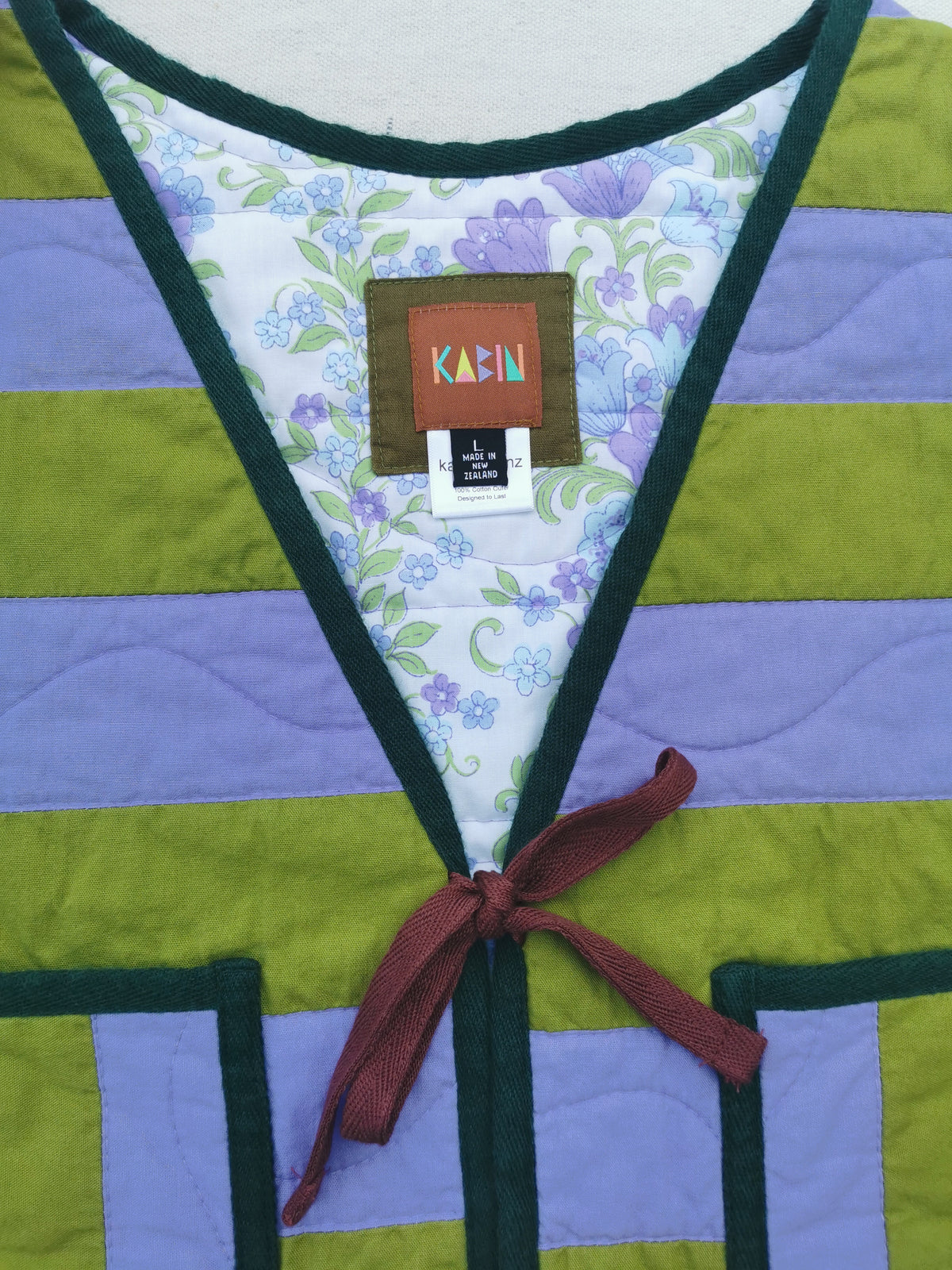 Close-up of a handmade quilted vest by KABIN with olive green and lavender horizontal stripes, dark green trim, a floral-patterned inner lining, and a dark red tie closure. A Kabin brand label is visible at the neckline.