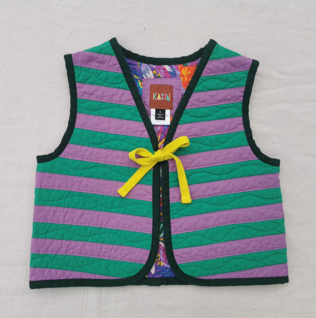 A handmade quilted children's vest by KABIN with horizontal purple and green stripes, dark green trim, and a yellow tie at the centre front. 