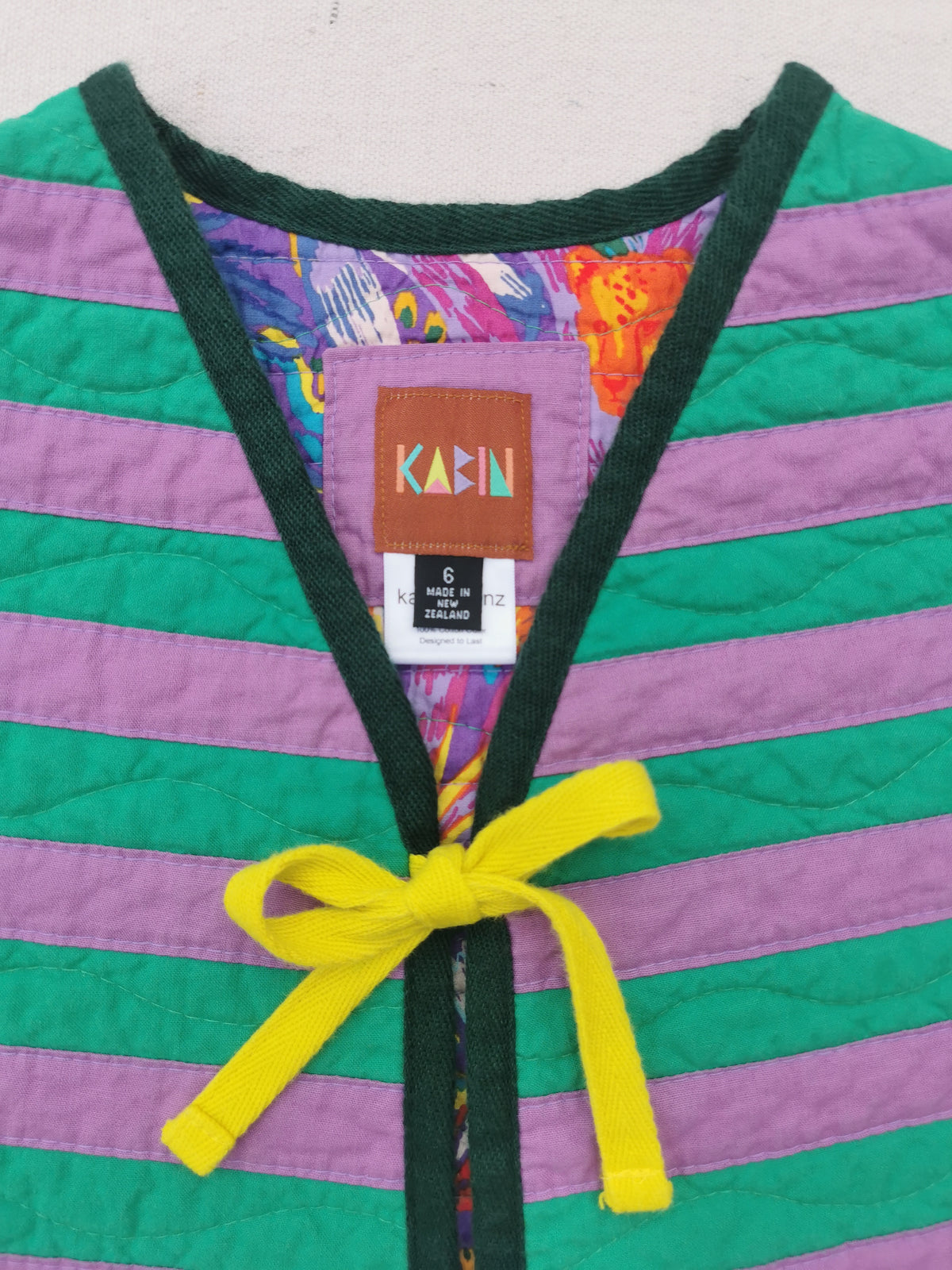 A close-up of a handmade quilted vest by KABIN, featuring horizontal stripes in green and purple with dark green trim and a bright yellow tie at the front. 