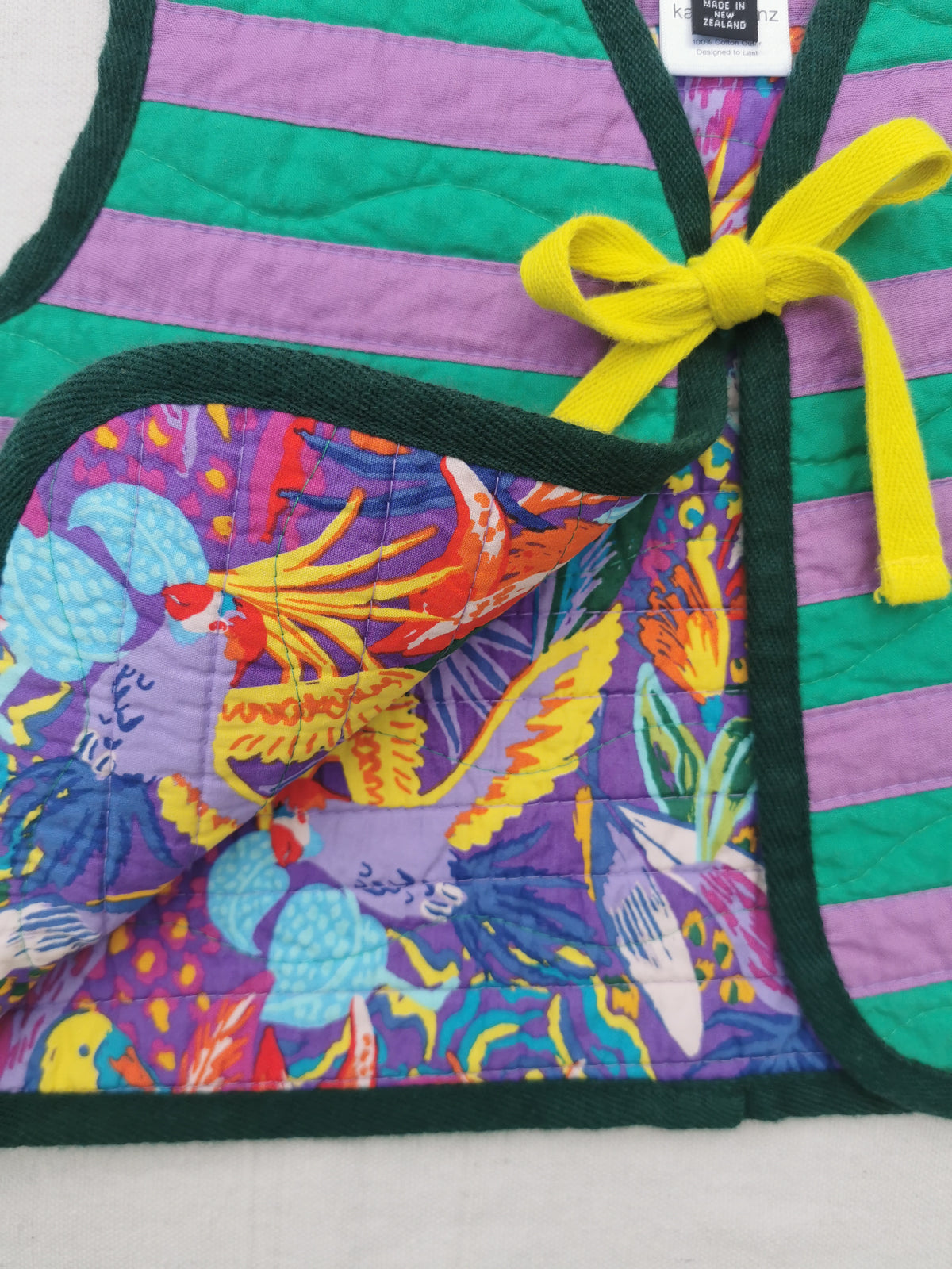 A close-up of a handmade quilted vest with dark green trim, purple and green horizontal stripes, a bright yellow tie, and a colourful floral-patterned lining by KABIN NZ.