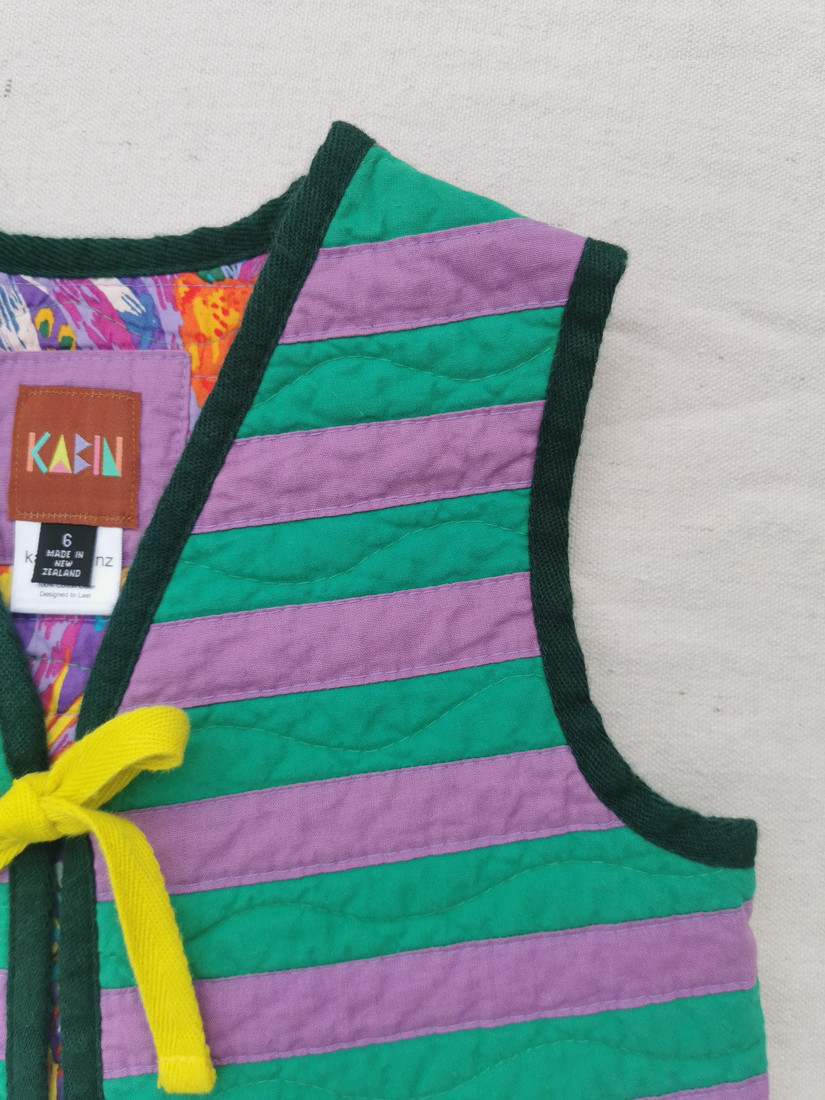 A close-up of a quilted vest with horizontal stripes in green and purple, dark green trim, a yellow tie closure, and a colourful interior lining. A brand label reading KABIN is visible.