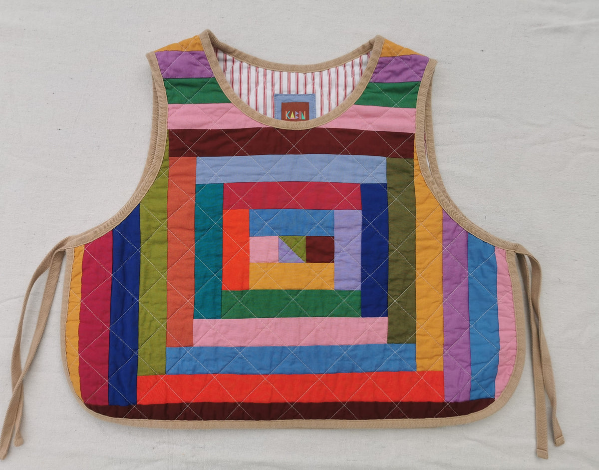 A handmade quilted vest with a colorful spiral log cabin patchwork pattern and beige tie straps, laid flat on a white fabric background by KABIN NZ.