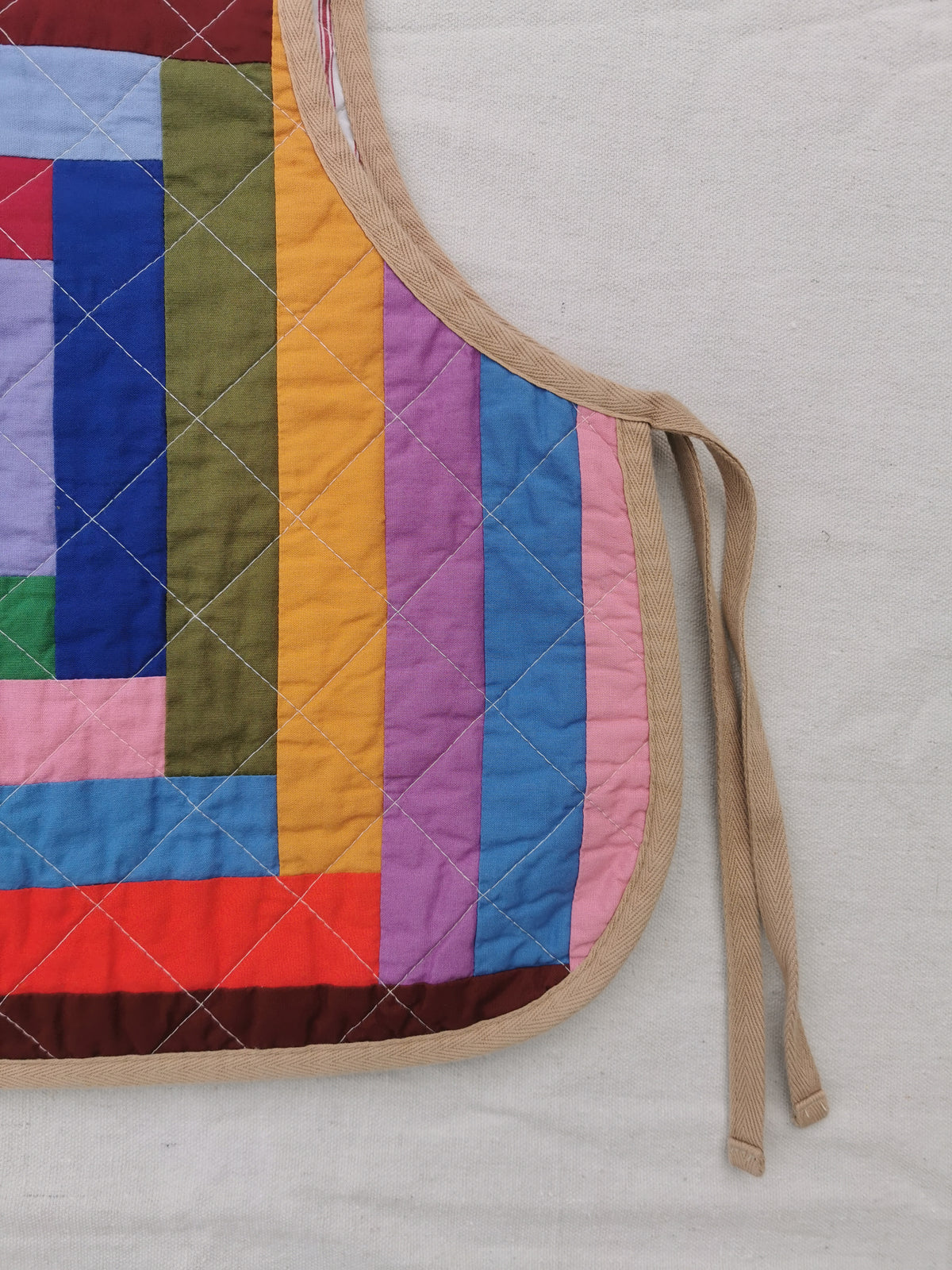 A close-up of a colourful quilted side tie vest with a geometric log cabin patchwork pattern and beige ties, laid flat on a light fabric background by KABIN NZ.