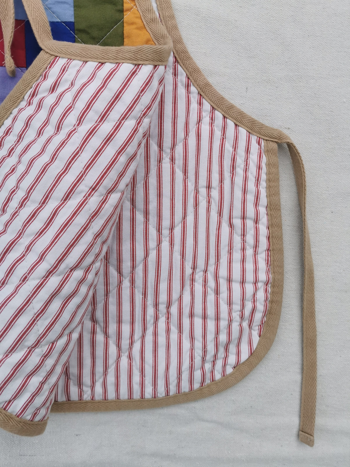 A close-up of a handmade quilted side tie vest with a white background, red and pink vertical stripes, and tan trim, laid flat on a light fabric surface by KABIN NZ.