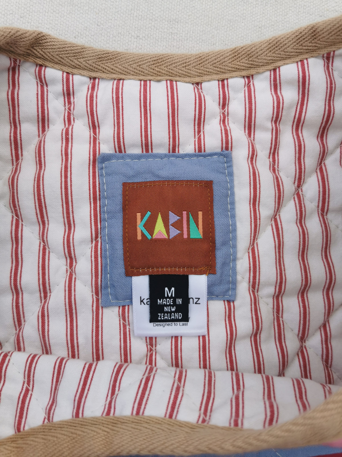 Close-up of a quilted vest showing a red and white striped interior, tan  trim, and a central sewn label with the brand name KABIN in colorful letters. Size medium, made in New Zealand.