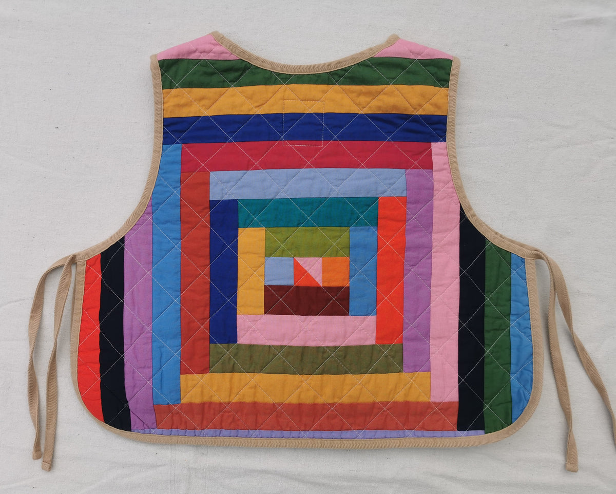 A handmade quilted side tie vest with a colorful patchwork spiral log cabin pattern, featuring beige ties and trim, laid flat on a white fabric background by KABIN NZ.