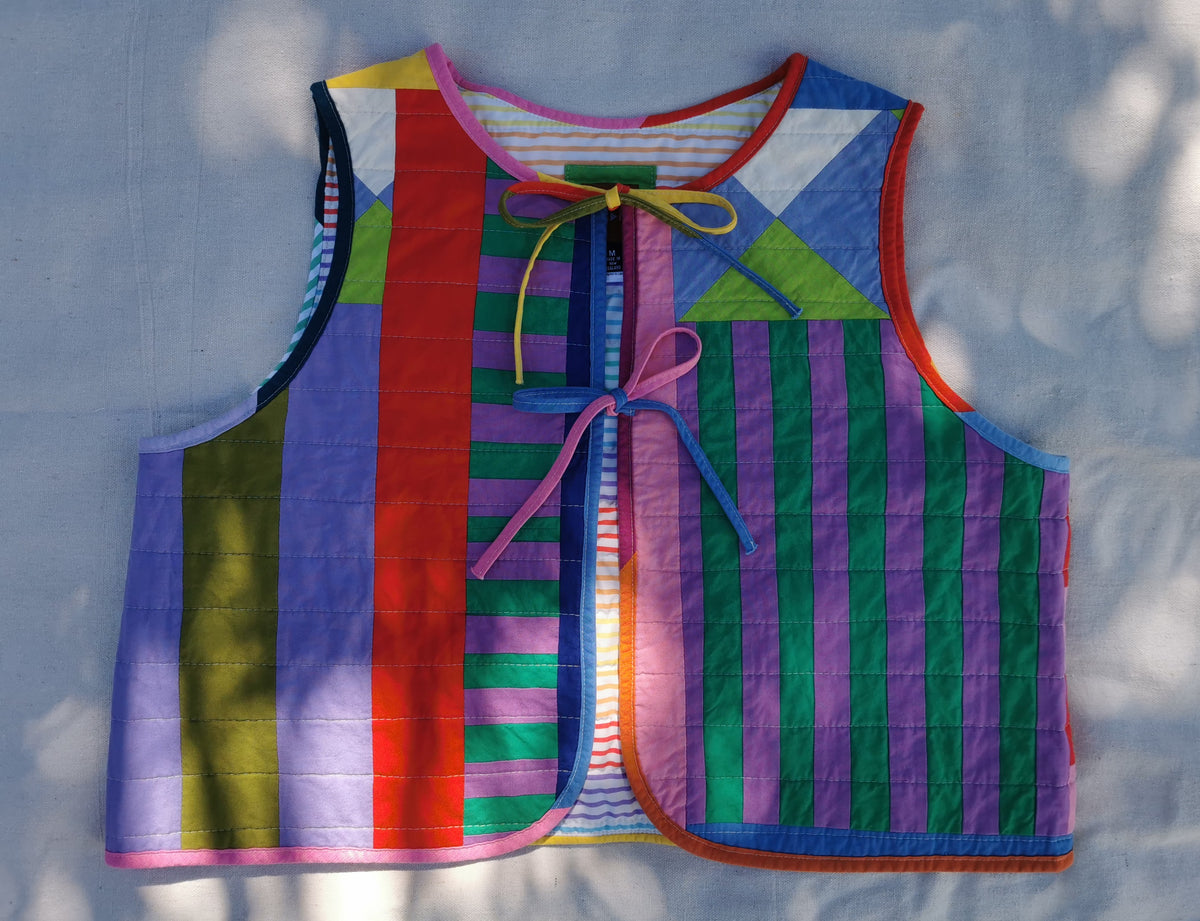 A colourful handmade quilted vest with a improv patchwork design featuring vertical stripes in purple, green, red, yellow, and blue, laid flat on a light surface with dappled sunlight by KABIN NZ.