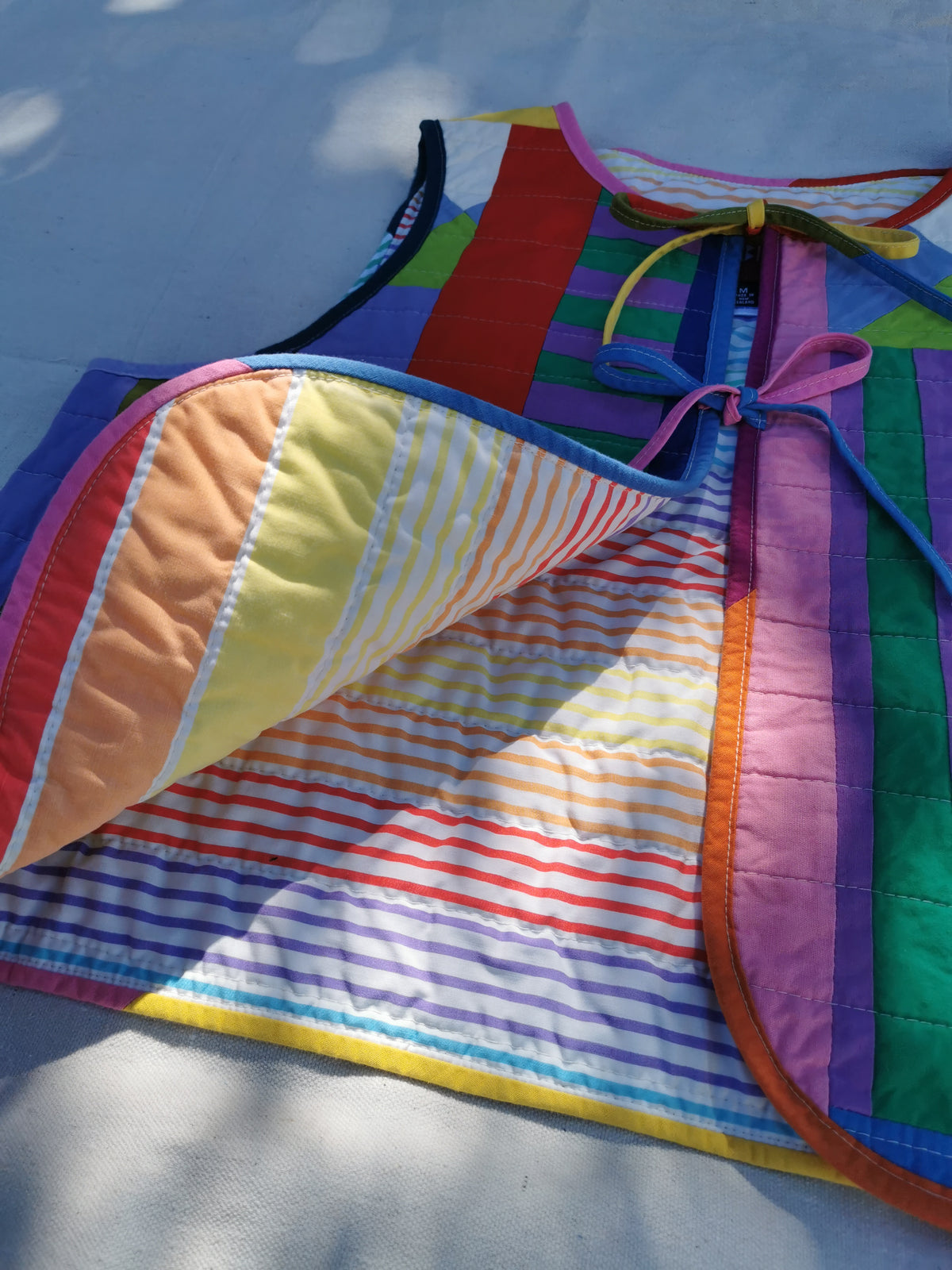 A colourful handmade quilted vest laid outdoors on a light surface, shown with one side folded over to reveal striped and solid fabric panels in rainbow colours including yellow, orange, red, blue, green, purple, and pink.