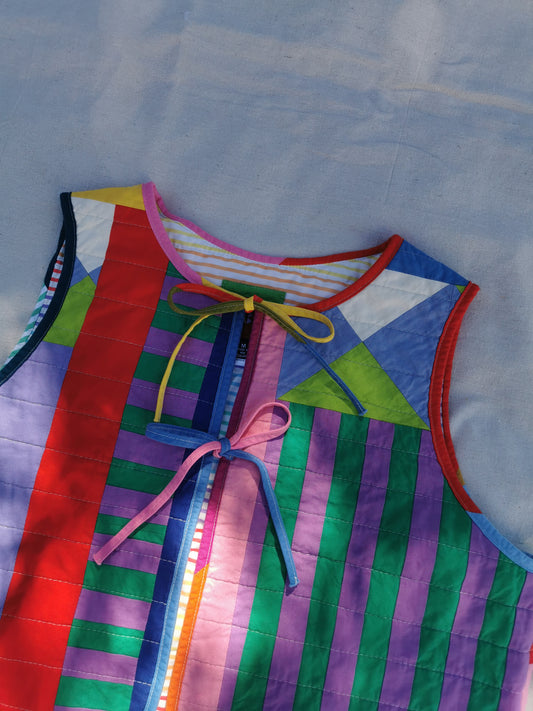 A colourful handmade quilted vest with vertical stripe panels in red, green, purple, blue, and pink, featuring tie closures at the neckline, laid flat on a light fabric background by KABIN NZ.