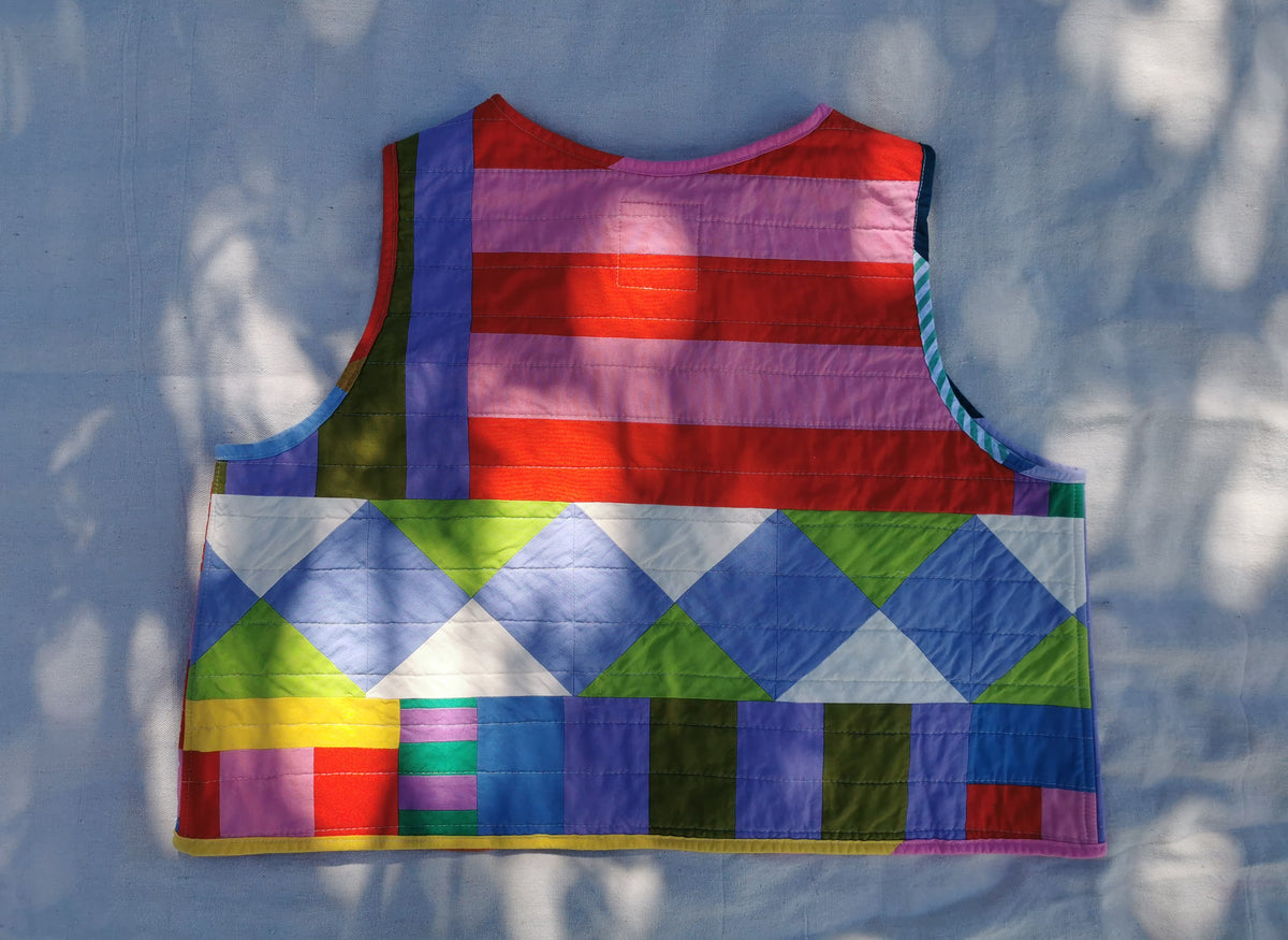 A colourful handmade quilted vest with a striped red, purple, and pink upper section and a geometric diamond pattern lower section, laid flat on light fabric background with dappled sunlight by KABIN NZ.