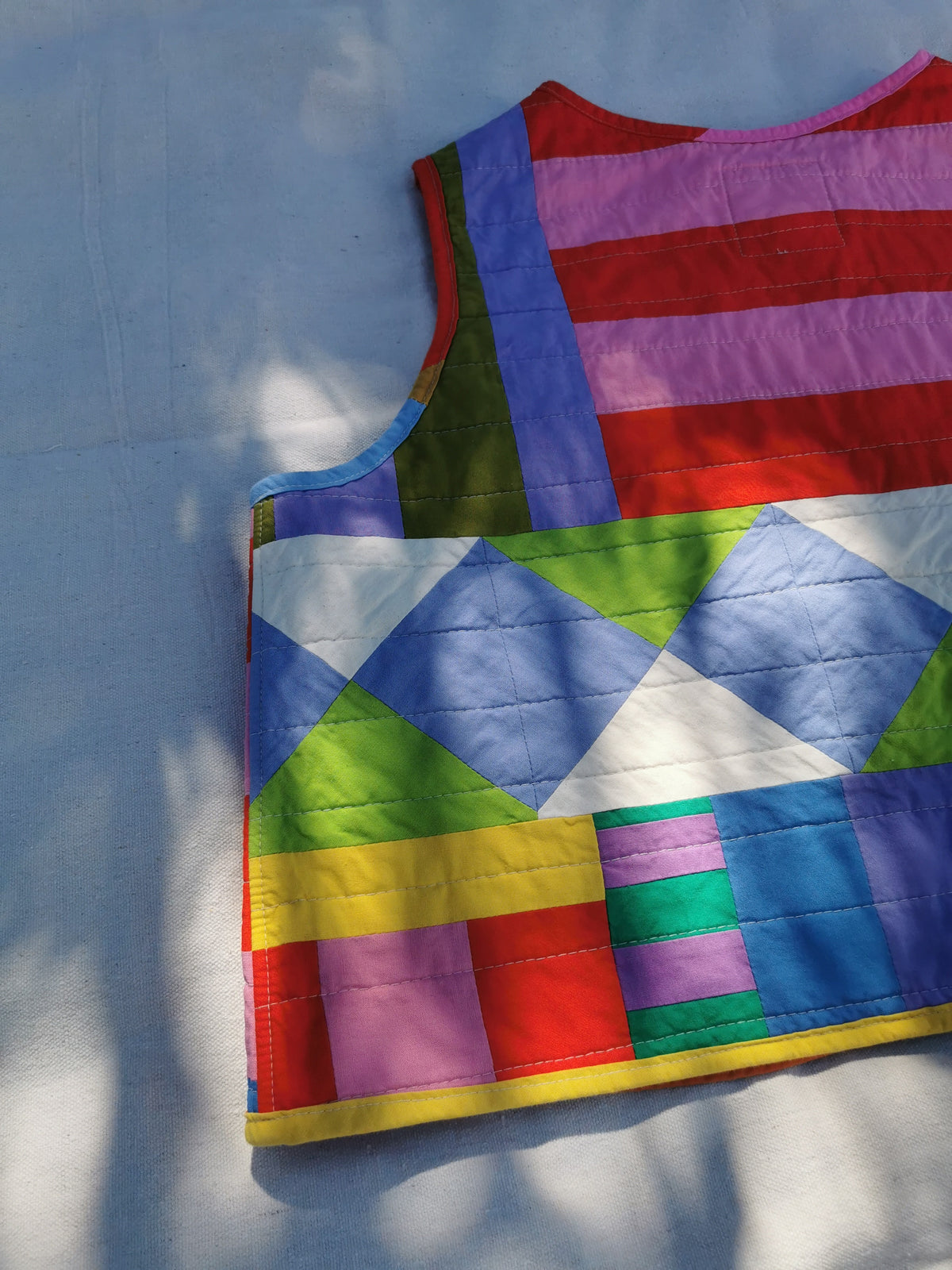 A colourful handmade quilted vest with a geometric patchwork pattern, laid flat on a light fabric background with dappled sunlight by KABIN NZ.