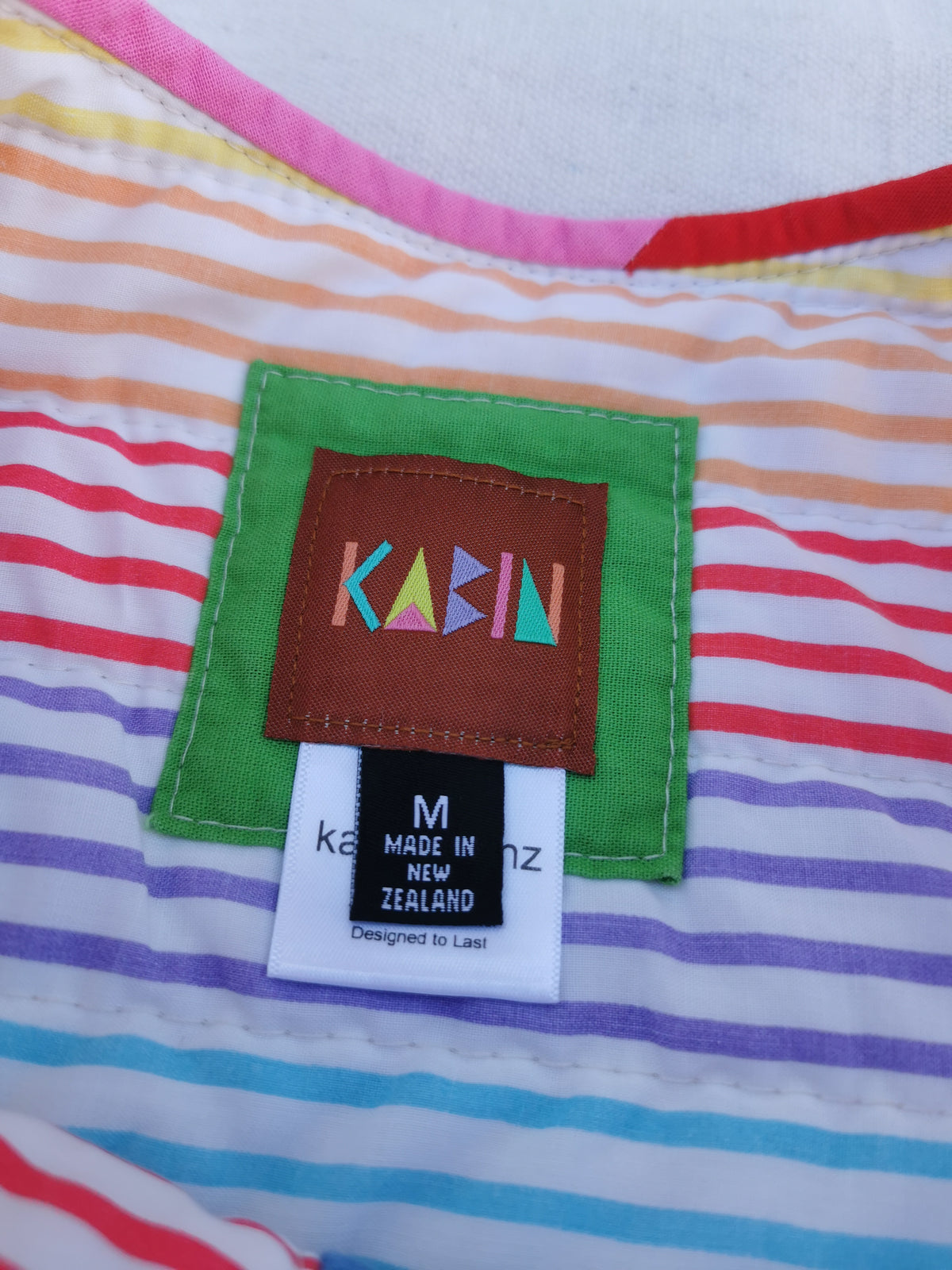 Close-up of a garment label patch reading KABIN, sewn onto a striped fabric with pink, orange, yellow, green, blue, and purple horizontal bands. Size medium, made in New Zealand.