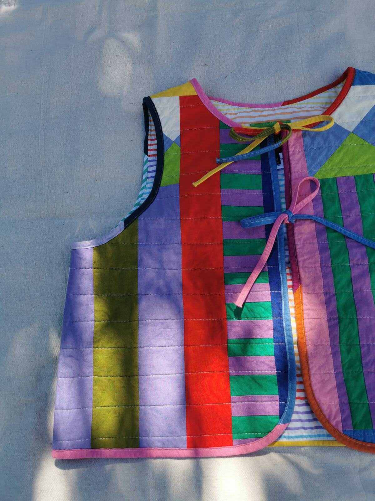 A handmade quilted vest with a colourful striped patchwork pattern, laid flat on a light fabric background with dappled sunlight by KABIN NZ.