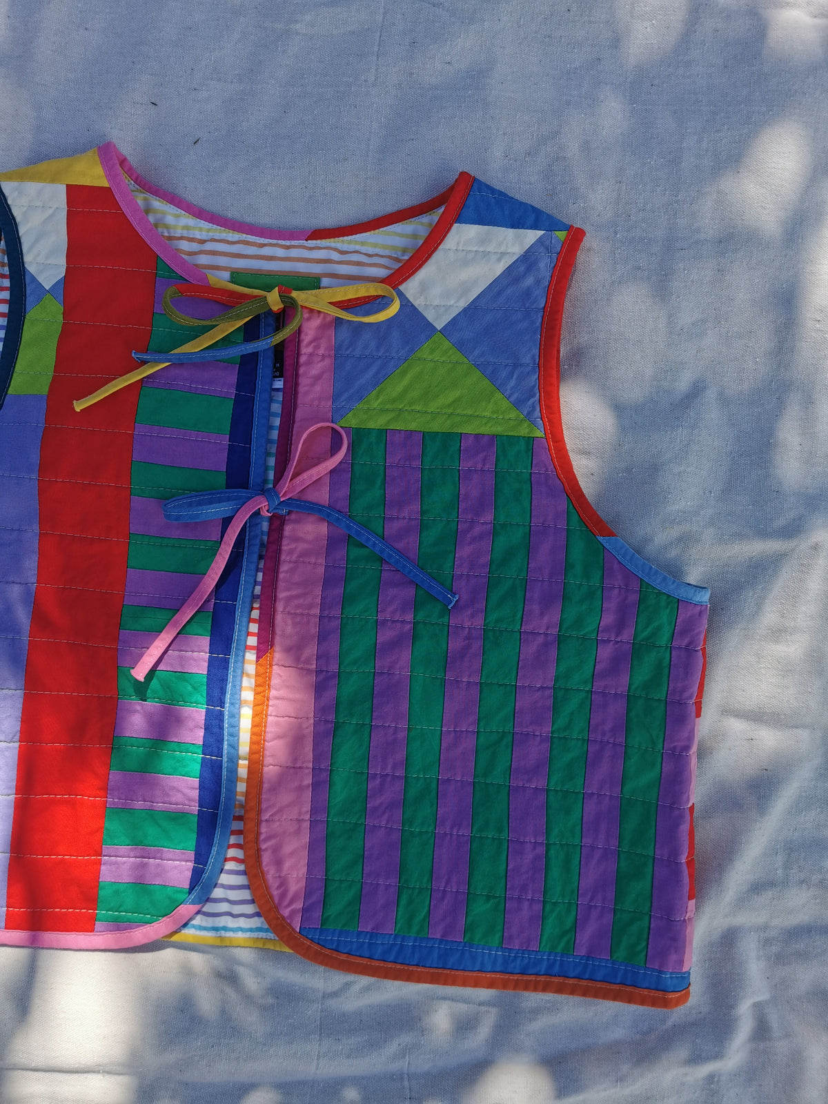A colourful handmade quilted vest with geometric and striped patterns, featuring tie closures, laid flat on a light fabric background with dappled sunlight.