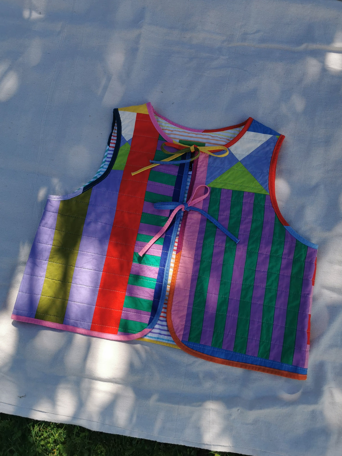 A handmade patchwork vest with colourful striped and geometric panels, laid flat on light fabric outdoors with dappled sunlight by KABIN NZ.