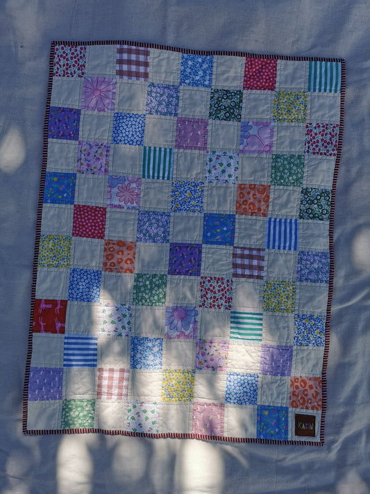 A handmade patchwork quilt laid flat on a light fabric background, featuring a grid of colorful patterned and striped square patches with a dark red and white striped border by KABIN NZ.