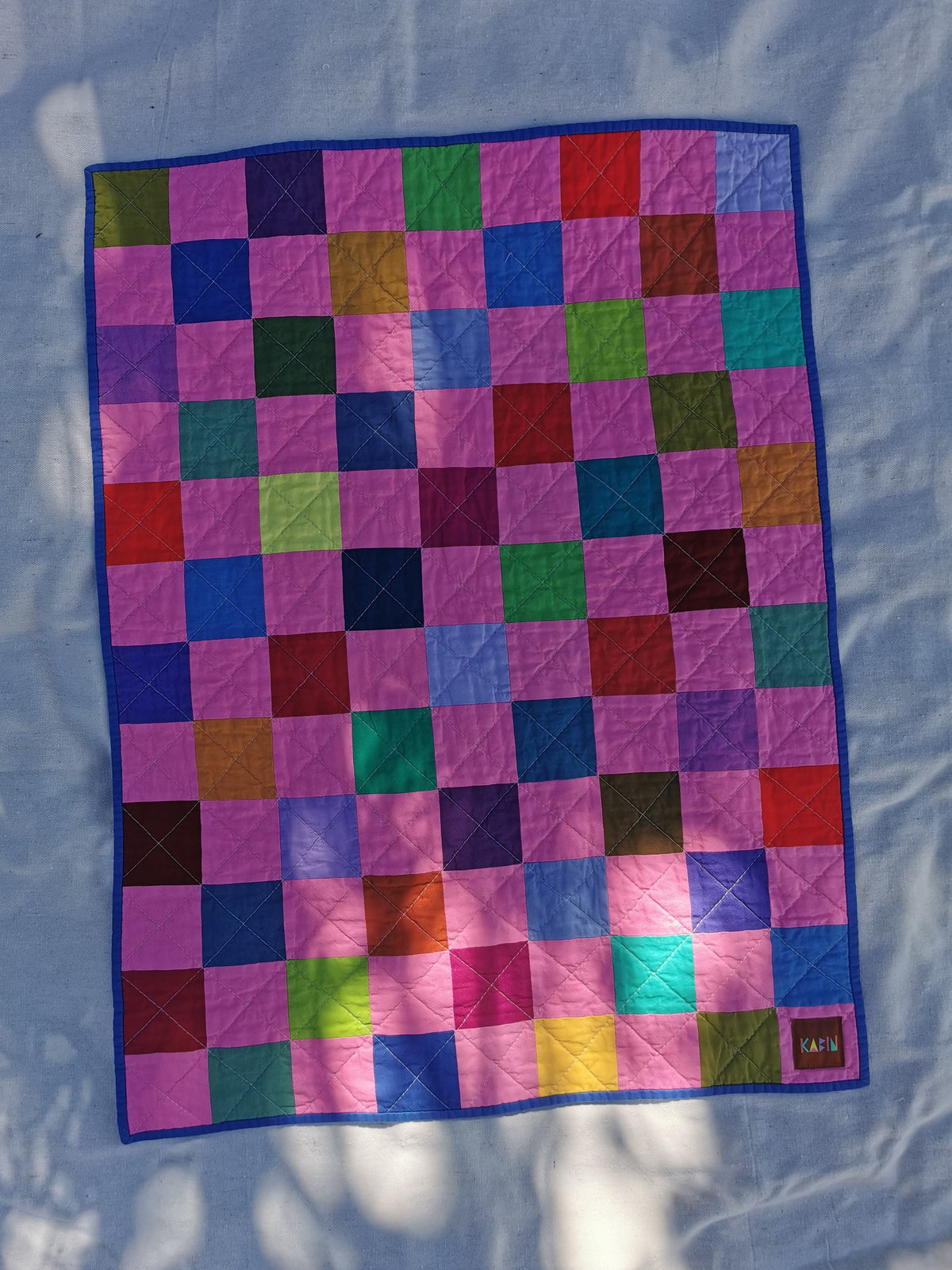 A colourful patchwork quilt with a grid pattern of bubblegum pink multi coloured squares, bordered in blue, laid flat on a light fabric background by KABIN NZ.