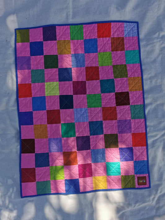 A colourful patchwork quilt with a grid pattern of bubblegum pink multi coloured squares, bordered in blue, laid flat on a light fabric background by KABIN NZ.