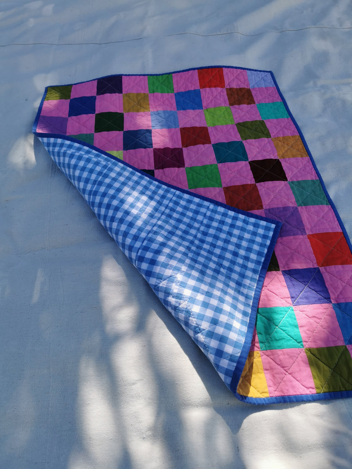 A handmade patchwork quilted blanket with a multicolor front and a blue-and-white gingham back, laid out on a light fabric surface with dappled sunlight by KABIN NZ.