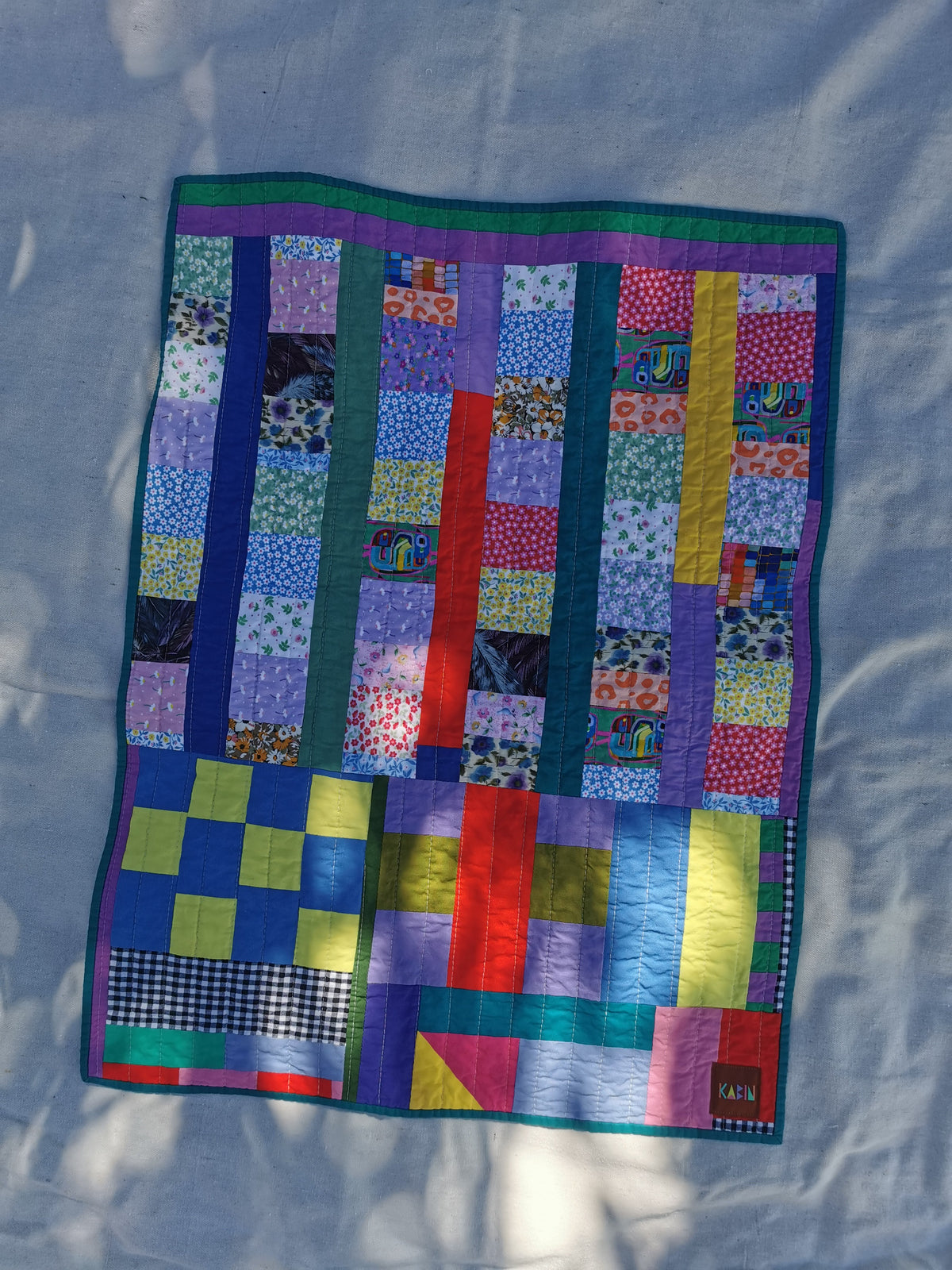 A colorful handmade patchwork quilt laid flat on a light fabric background with dappled sunlight by KABIN NZ.