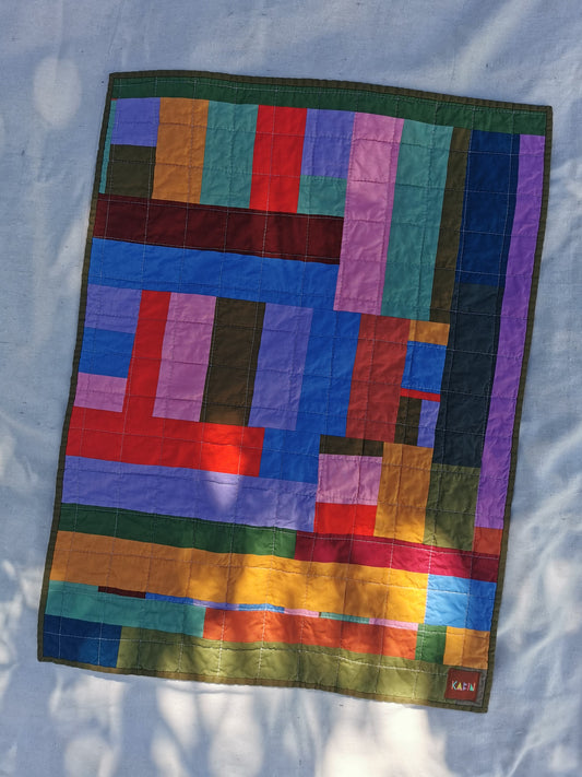 A colorful handmade patchwork quilt  by KABIN NZ with a geometric block pattern, laid flat on a light blue fabric background with dappled sunlight.