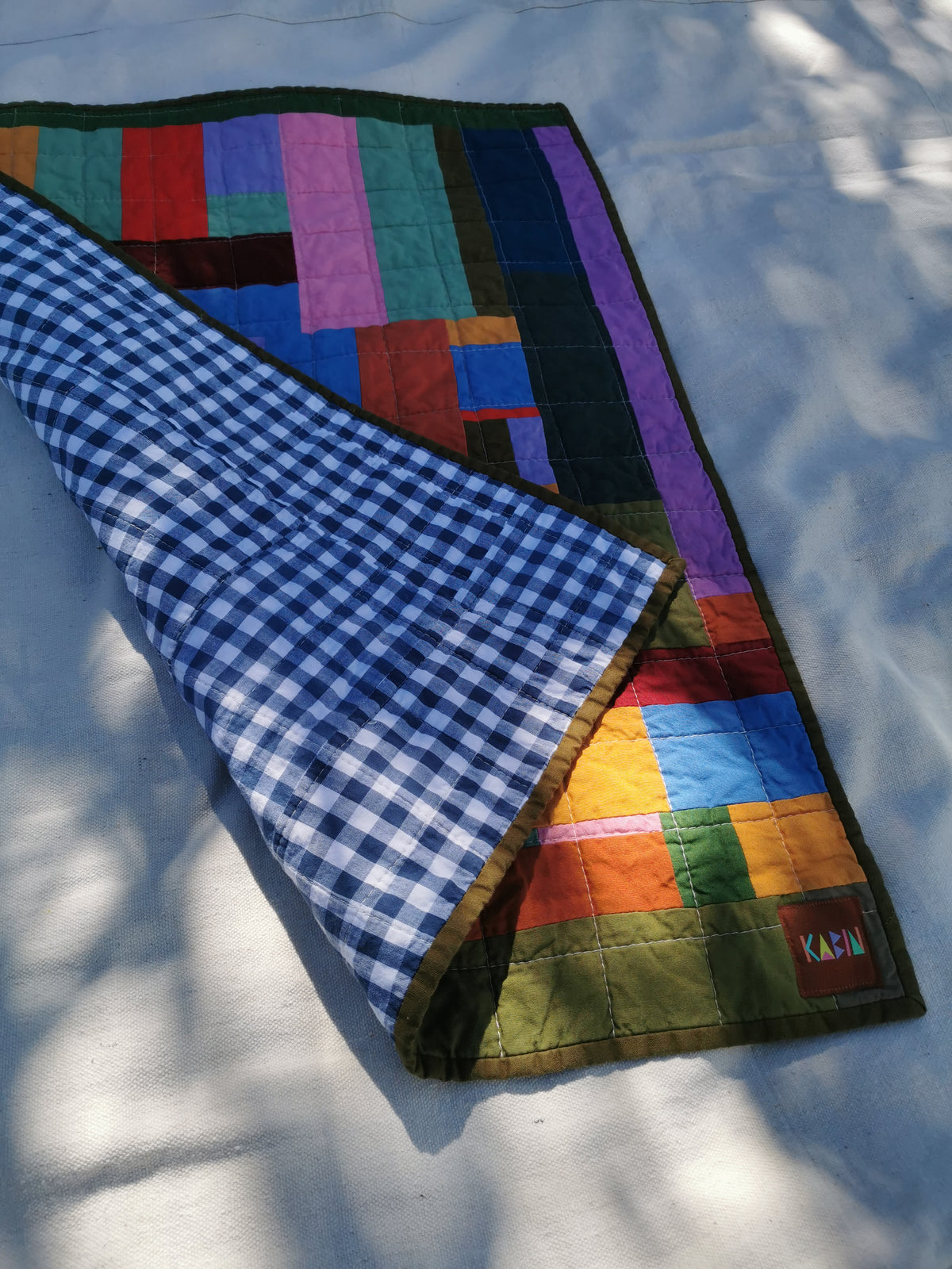 A folded quilted blanket with a multicolor patchwork design and a blue-and-white gingham reverse side, laid on a white surface with dappled sunlight and shadows by KABIN NZ.