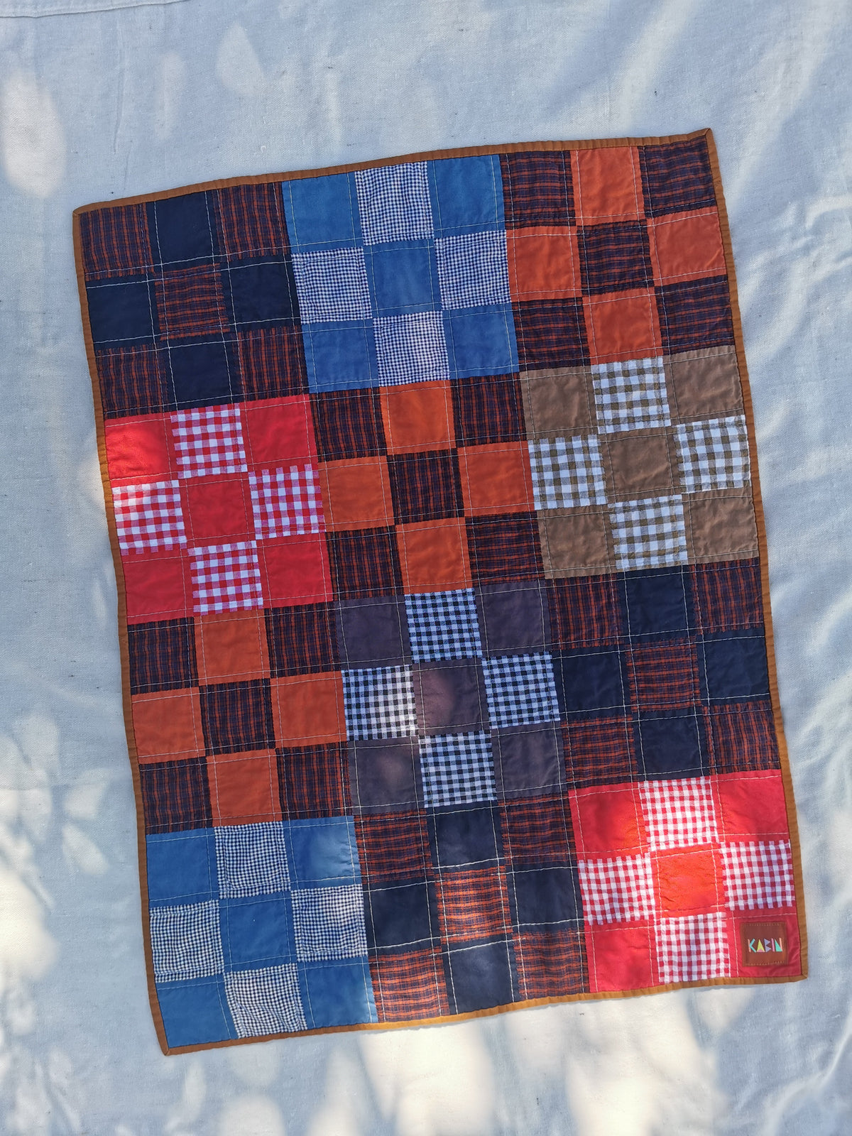 A handmade patchwork quilt laid flat on a light fabric background. The quilt features a grid pattern of various patchwork squares in blue, red, orange, brown, and navy, with some gingham and plaid designs by KABIN NZ.
