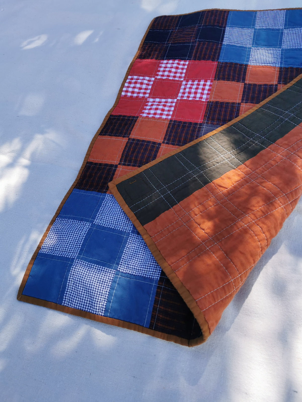A handmade patchwork quilt laid out on a light surface with dappled sunlight. The quilt features a mix of blue, red, brown, and black checkered and gingham patterns, with a solid brown border by KABIN NZ.
