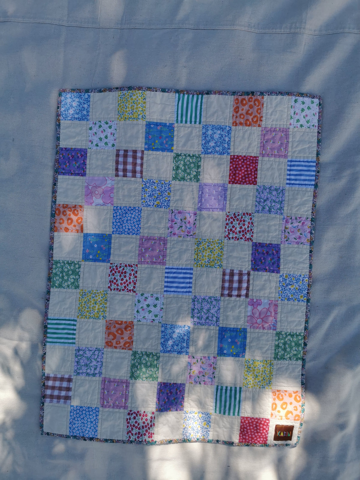 A square patchwork quilt laid flat on a light fabric background. The quilt features a grid of colourful square patches in various patterns including stripes, florals, plaid, and solid colors, with a dark border by KABIN NZ.