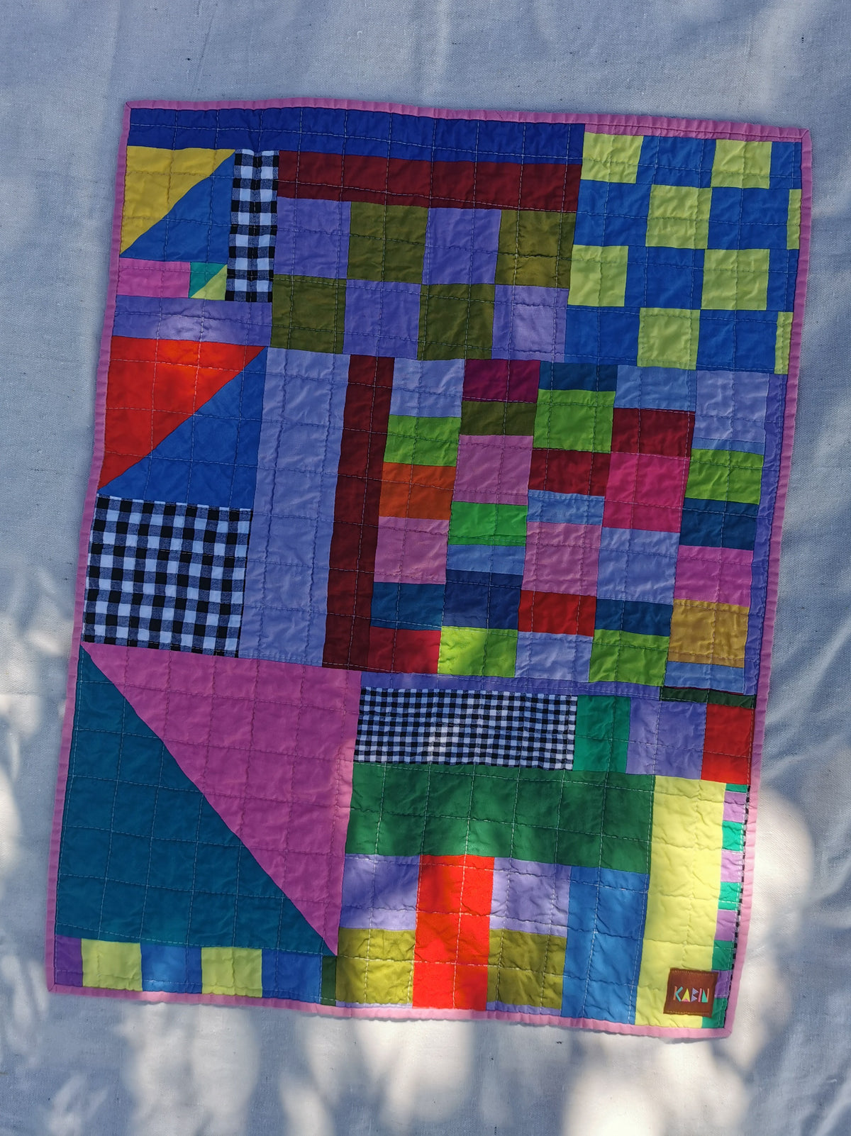 A KABIN NZ colourful geometric handmade patchwork quilt laid flat on a light fabric background. The quilt features a variety of colored blocks, including pink, blue, green, yellow, red, and purple, with some gingham patterned sections and a pink border.
