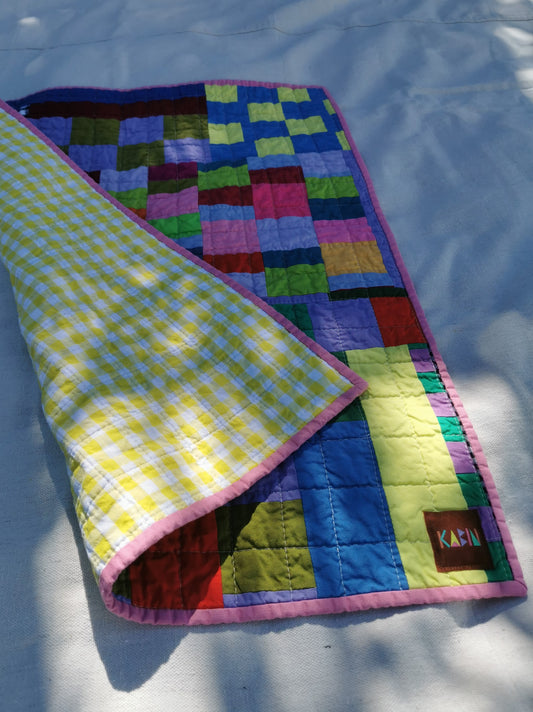 A colourful patchwork quilt by KABIN NZ with a yellow and white gingham reverse side, laid out on a white fabric background with dappled sunlight and shadows. A small brown brand patch reading 'KABIN' is visible on the quilt.