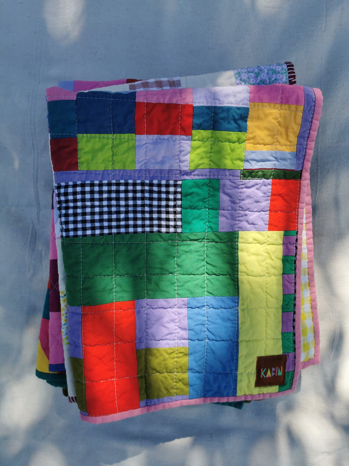 A colourful patchwork quilted blanket with a pink border and a small brown 'KABIN' label, draped against a light fabric backdrop with dappled sunlight.