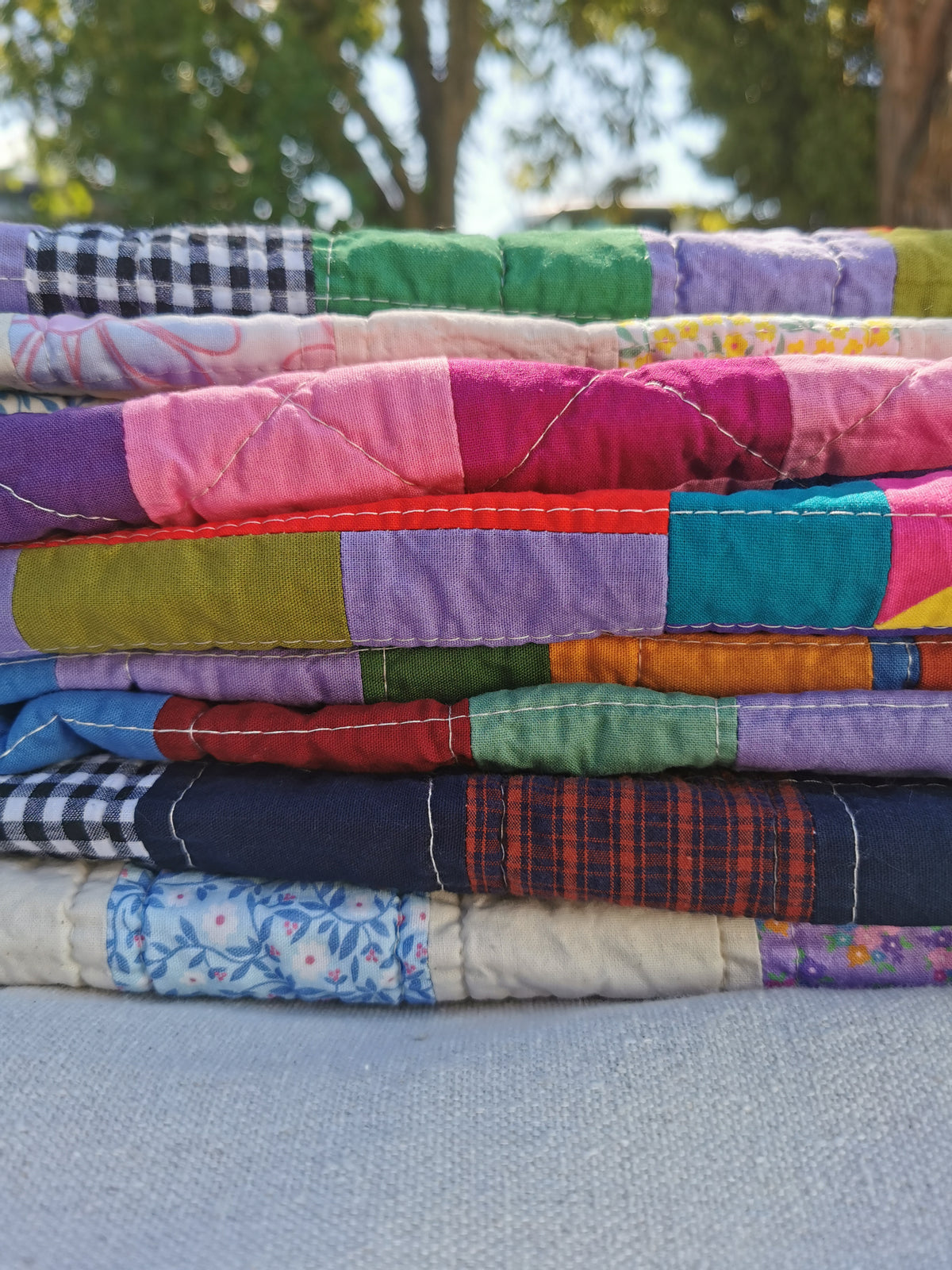 A stack of colourful handmade patchwork quilts by KABIN NZ, with various patterns and vibrant colors, photographed outdoors on a light gray fabric surface with blurred green trees in the background.