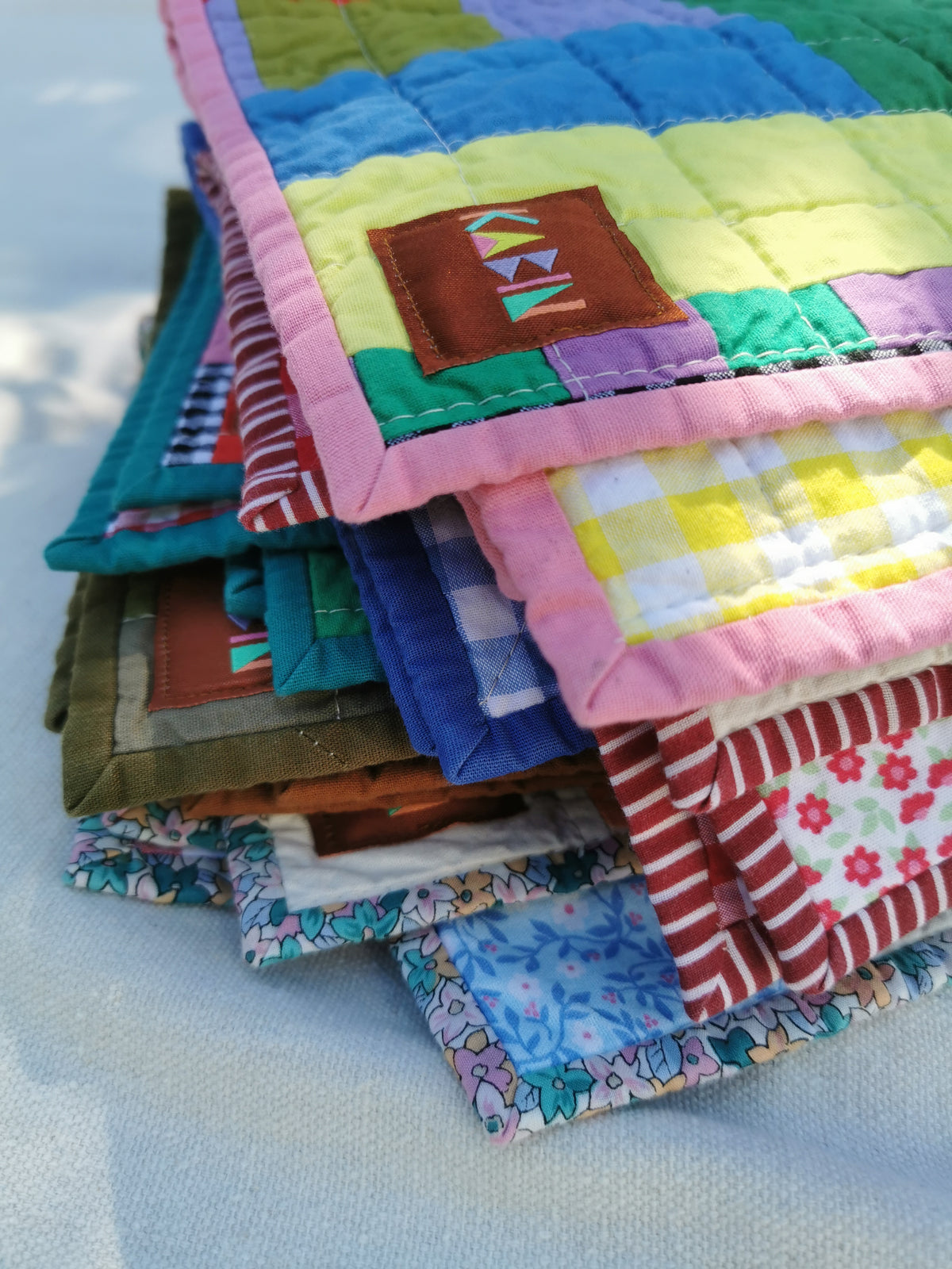 A stack of colorful handmade quilted fabric small quilts with patchwork panels, floral prints, and pink borders, photographed outdoors on a light fabric surface by KABIN NZ.